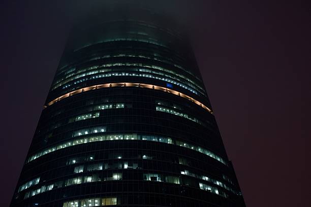 A modern skyscraper illuminated at night shrouded in fog, showcasing its sleek architecture and glowing windows.