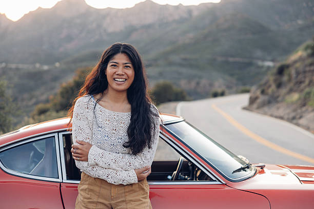 Woman smiling while leaning on the car during a road trip traveling Southern California with a vintage car on coastal winding roads.