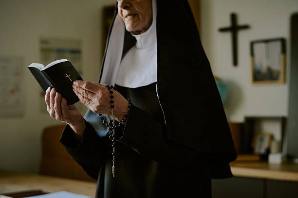 Elderly nun standing in classroom while reading bible and holding rosary beads in hands. Crucifix visible on wall behind her complementing tranquil setting