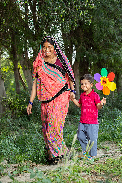 Smiling boy holding pinwheel toy walking with mid adult mother dressed in sari on lush agricultural landscape