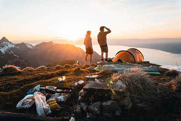 View past campfire and food to young men enjoying sunrise near tent on mountain peak above lake Geneva and distant mountains, Mount Grammont, Valais, Swiss Alps