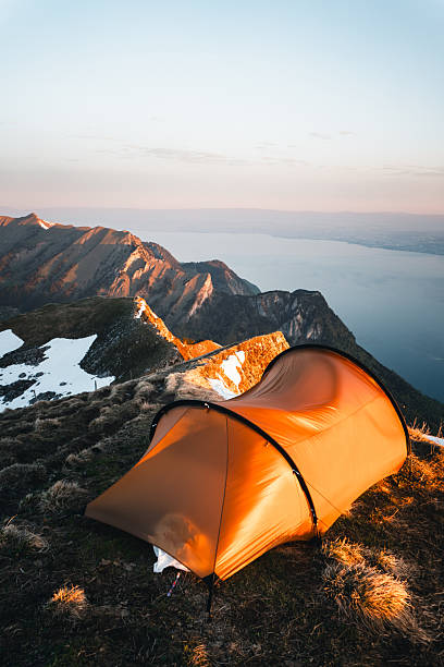 Illuminated tent at campsite on mountain peak above lake Geneva and distant mountains, Mount Grammont, Valais, Swiss Alps