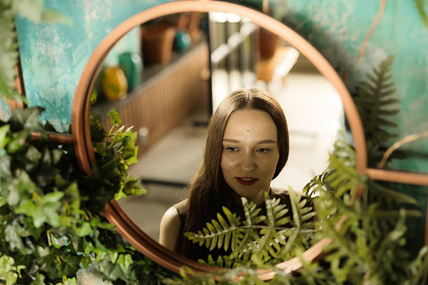 A woman gazes thoughtfully into a large mirror, surrounded by vibrant and lush greenery in a serene setting