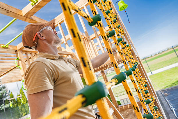 A construction worker examines a bright yellow climbing structure at a playground construction site. He is wearing a tan cap and safety glasses while focusing on the sturdy ropes. Surrounding him is an open field under a clear blue sky, indicating the project's progress during the daytime.