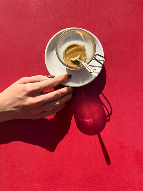 Woman hand with dark nail polish holding an empty glass espresso coffee cup with a spoon on a red table in natural light, coffee break concept with copy space