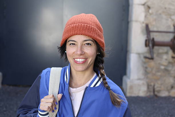 A very beautiful young female student in her forties is walking on the street with her backpack. She wears an orange beanie and a classic university jacket.