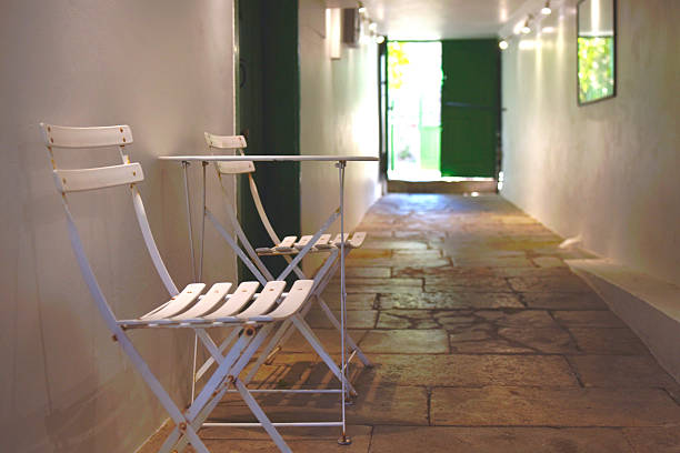 A long, narrow corridor with a slight incline, leading towards a dimly lit doorway. A solitary chair sits in the distance, adding a touch of mystery to the scene.