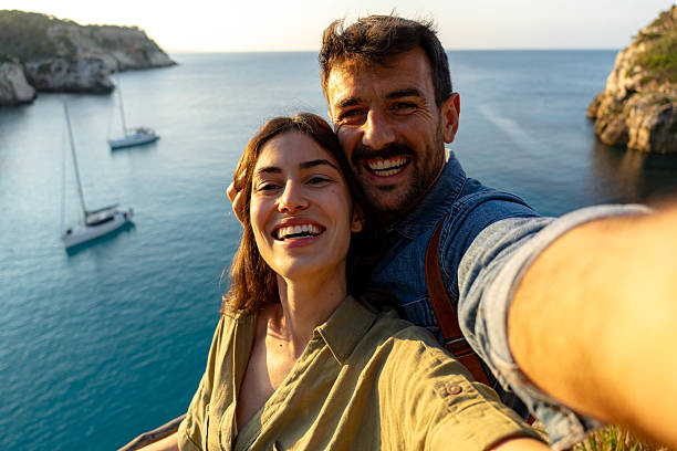Smiling Couple Enjoying Scenic Ocean View Happy couple taking a selfie with a stunning ocean backdrop featuring sailboats and cliffs, capturing a memorable travel moment yacht selfie stock pictures, royalty-free photos & images