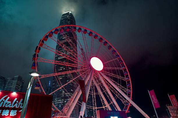 The Hong Kong Observation Wheel glows in vivid red against a dramatic, cloud-covered sky, with the towering International Finance Centre (IFC) skyscraper looming behind. This striking night shot captures the vibrant energy of the Central Harbourfront district, where the iconic ferris wheel serves as a centerpiece of leisure and city life. The mist, light beams, and towering architecture together create a cinematic atmosphere unique to Hong Kong’s skyline.