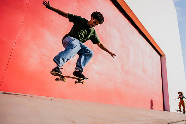 Young person performing a skateboard trick in mid-air, in front of a large red wall in an urban setting. Wearing a green t-shirt and loose jeans. The scene takes place outdoors under strong natural light. In the background, a second person is skateboarding at Ibirapuera Park in São Paulo, Brazil