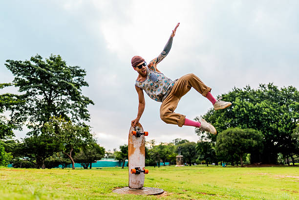 Mid adult man performing an extreme skateboard trick in a park, balancing with one hand on the upright skateboard while airborne. Wearing a printed tank top, beige pants, pink socks, and a cap. The scene takes place on a grassy area with trees and a cloudy sky in the background