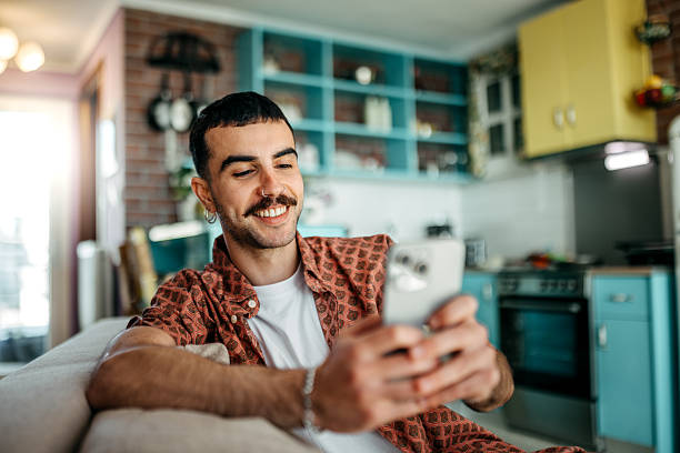 istock Cheerful hipster man using smart phone while relaxing on a sofa at home 2215969662