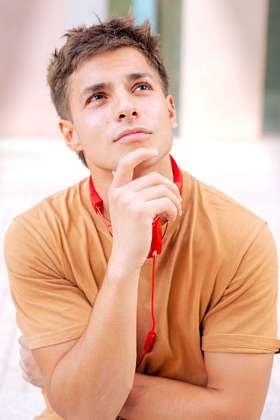Young man wearing casual clothes and red headphones, looking up thoughtfully in a bright outdoor setting. Contemplative expression and modern style.