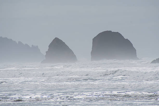 Tall, iconic sea stacks, including the famous Haystack Rock, rise dramatically against a cloudy sky at Cannon Beach in Oregon, on April 12, 2025. These rugged rock formations are a hallmark of the Pacific Northwest coastline and a popular natural landmark.