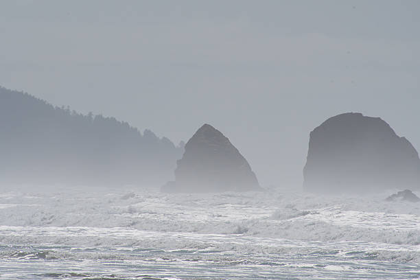 Tall, iconic sea stacks, including the famous Haystack Rock, rise dramatically against a cloudy sky at Cannon Beach in Oregon, on April 12, 2025. These rugged rock formations are a hallmark of the Pacific Northwest coastline and a popular natural landmark.