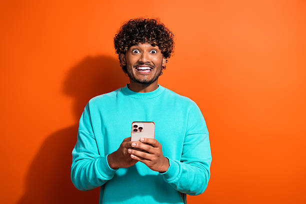 istock Photo of young surprised funny positive man indian tik tok user wearing teal pullover using smartphone isolated on orange color background 2215975477