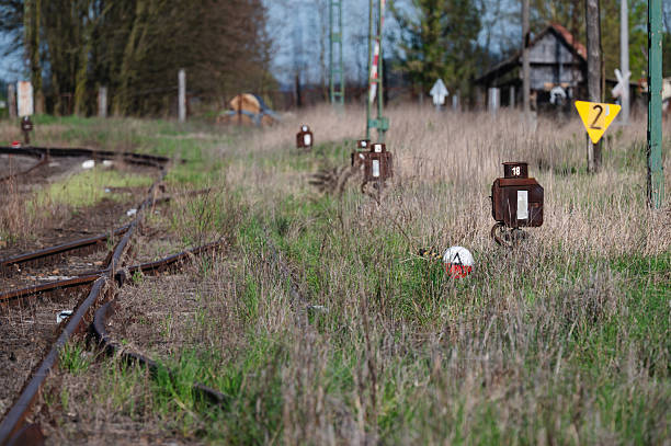 An old railway track runs through an overgrown grassy field, with multiple rusted switch indicators and signal markers visible. The scene conveys a sense of abandonment and rural decay, with weathered infrastructure and an unused track diverging into the distance.