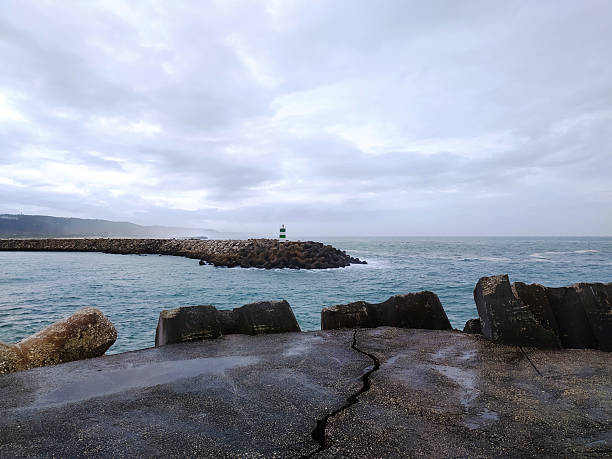 Gloomy seascape featuring a breakwater leading to a distant lighthouse. Concrete structures frame the scene, creating depth. Perfect for themes of resilience and guidance.