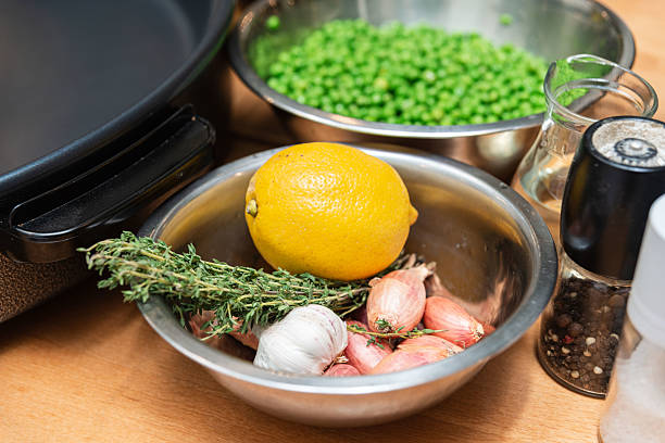Stainless steel bowl filled with fresh lemon, shallots, garlic, and thyme, next to peas and seasoning on a kitchen counter.