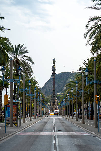 View of the Christopher Columbus Monument with the World Trade Center building in the background, seen from the waterfront in Barcelona, Spain. The image captures the iconic statue pointing towards the sea, framed by modern architecture under a soft sky.