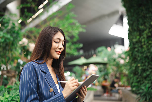 istock Young woman thoughtfully sketches notes or ideas on her tablet amidst lush greenery, blending innovation with tranquility in a vibrant urban setting. 2216007760