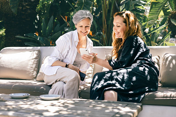 istock Two mature women smiling and looking at smartphone together while sitting outdoors on a terrace 2216007947