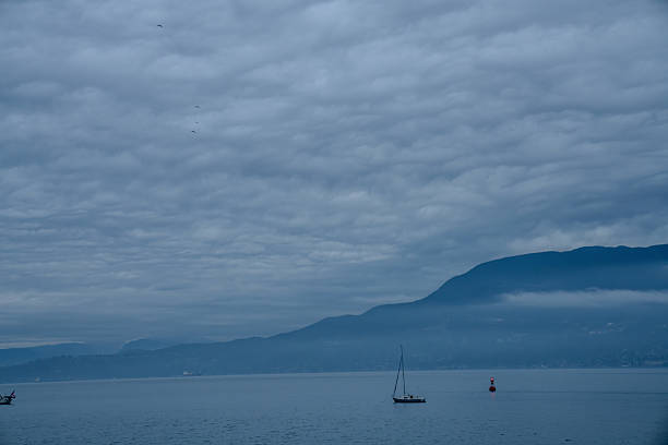 This tranquil image showcases a peaceful seascape under a textured, overcast sky. The dense clouds blanket the scene in soft gray and blue hues, creating a calm and subdued atmosphere. In the center, a lone sailboat drifts on the still water, evoking a sense of solitude and quiet reflection. To the right, a red buoy adds a subtle pop of color, breaking the otherwise muted palette. The background features a mist-shrouded mountain range, its silhouette fading gently into the horizon. The contrast between the serene water, the dramatic clouds, and the distant mountains creates a harmonious balance, capturing the beauty of nature in its simplest form.