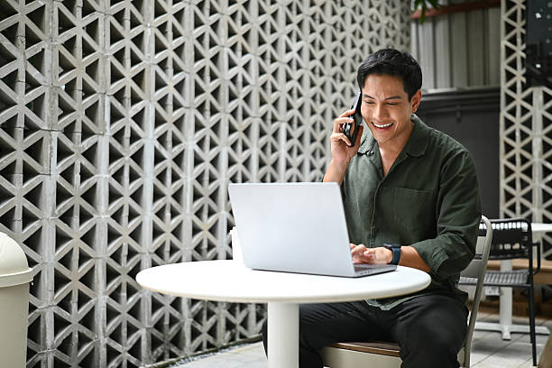 istock A man is sitting at a table with a laptop and talking on a cell phone. He is smiling while talking on the phone. 2216008927