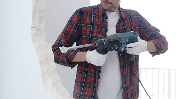 istock Male construction worker wearing red checkered shirt and protective white gloves is demolishing white wall with rotary hammer drill and generating dust during renovation project, closeup view 2216015739