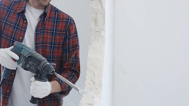 istock Male construction worker wearing red checkered shirt and protective white gloves is demolishing white wall with rotary hammer drill and generating dust during renovation project, closeup view 2216016629