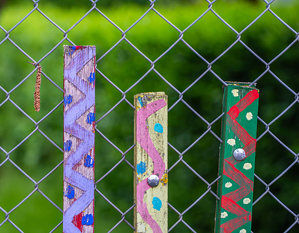Colorful Painted Wooden Posts on Chain-Link Fence Colorful painted wooden posts attached to a chain-link fence, with a blurred green background. The posts feature zigzag and dot patterns in vibrant colors. wood and wire fence designs pictures stock pictures, royalty-free photos & images
