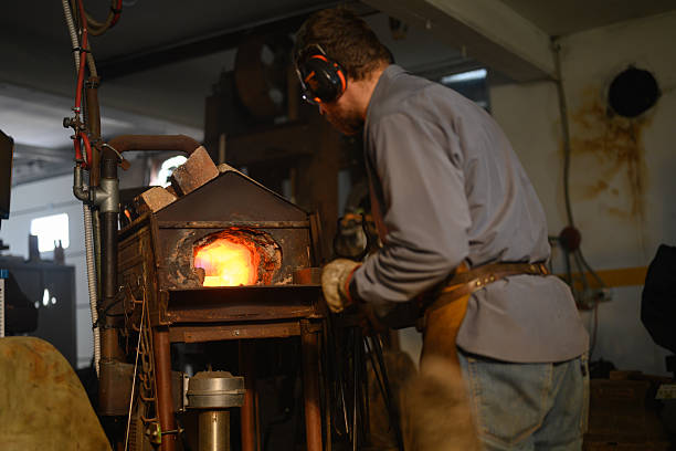 Side-angle view of a mid adult craftsman in protective gear working at a traditional forge, manipulating metal within the glowing furnace in an industrial workshop environment.