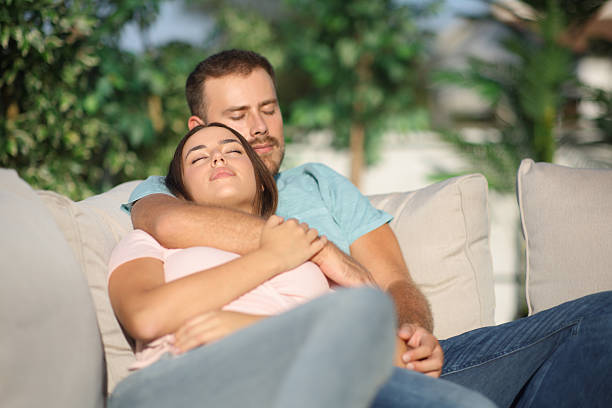 A man and woman relax on a grey sofa in a modern living room, one leaning back with eyes closed, the other casually seated. A remote sits nearby, suggesting a quiet movie night.