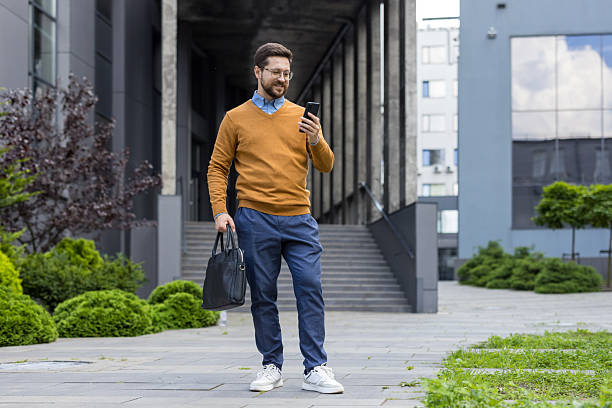 istock Young successful male businessman standing near office center with document bag and using mobile phone 2216043947