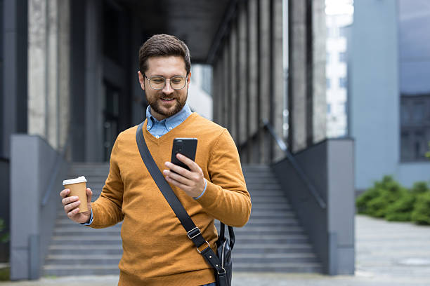 A young male office worker is standing outside a building, holding a cup of coffee and using a mobile phone with a smile