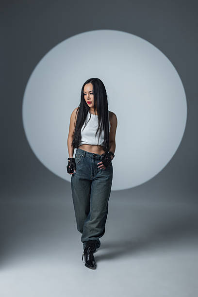 istock Confident young woman in a white crop top, jeans, and boots posing in a studio with dramatic lighting and a circular spotlight backdrop. Fashion, attitude, and style concept. 2216044302