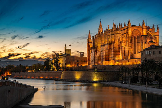 The majestic La Seu Cathedral in Palma de Mallorca glows warmly at dusk, its Gothic architecture reflected in tranquil waters under a vibrant twilight sky.