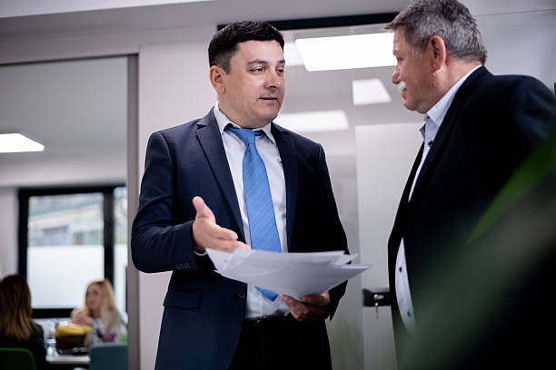 istock Two men in business attire discussing documents in an office environment with colleagues working in the background 2216058580