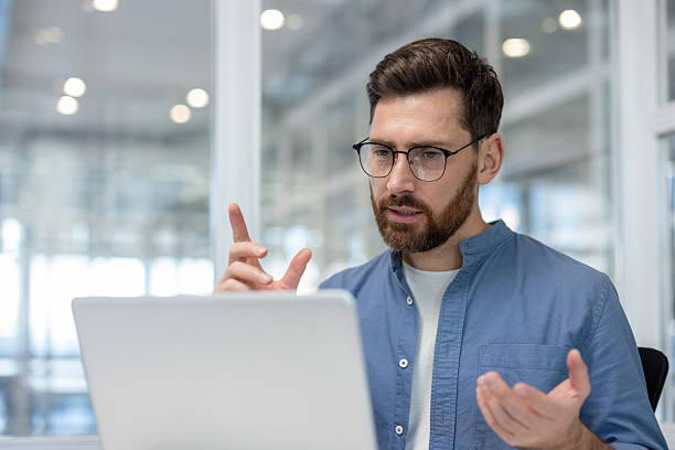 istock Serious concentrated man talking to colleagues and partners using laptop and video call. Close up of company employee working remotely inside office, serious business meeting 2216059449