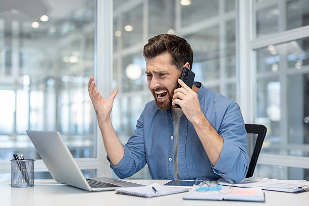 istock Angry man shouting while talking on mobile phone inside office. Company employee at workplace working on laptop, unhappy with conversation 2216059508