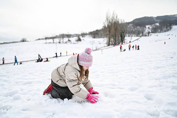 Girl making a snowman in a snowy park while other people are sledding