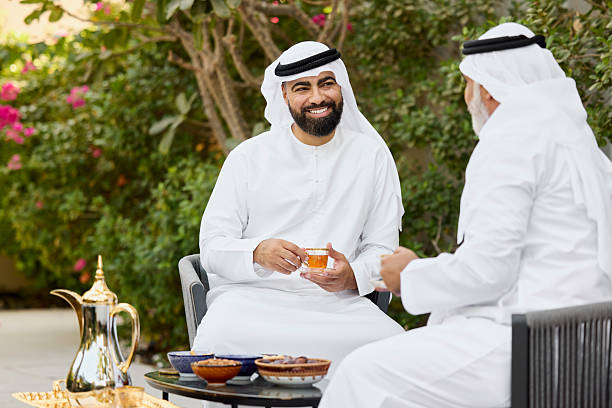 Smiling middle eastern man having tea with family member in back yard. Arab males are having conversation. They are wearing traditional attire.