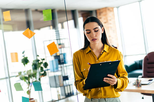 istock Young businesswoman in a modern office environment analyzing data on a clipboard surrounded by colorful sticky notes on a glass wall 2216065954