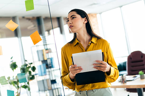 istock Confident businesswoman reviewing documents in a bright, modern office environment, thoughtfully analyzing sticky notes on a glass wall 2216066009