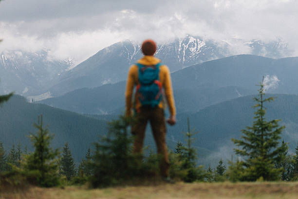 The unrecognisable male hiker looking at the majestic mountains view covered by snow and clouds in spring