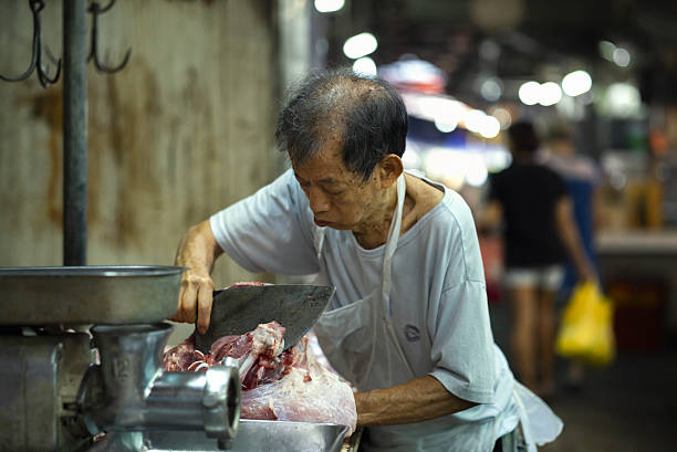 An elderly butcher concentrates on cutting fresh meat at a traditional wet market, embodying decades of dedication and craftsmanship. The worn walls and soft lighting emphasize the authenticity and everyday rhythm of local life.