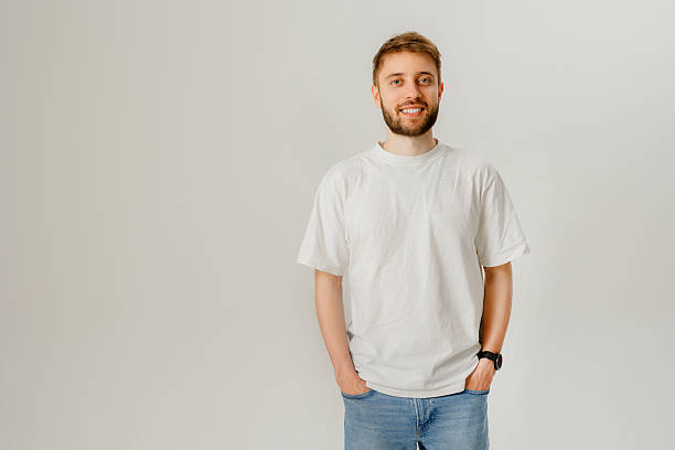 istock Young man stands casually in a simple white t-shirt with hands in pockets, smiling against a neutral background 2216094549