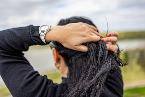 A close-up image of a woman tying her long black hair outdoors with both hands. She is wearing a black sweater and a silver watch, standing against a blurred natural background with cloudy skies and water in the distance. Selective focus