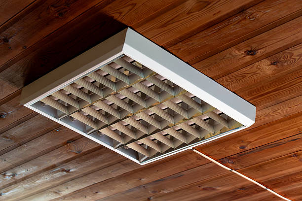 A fluorescent ceiling light fixture with a metallic grid is mounted on a wooden paneled ceiling, viewed from below in a rustic indoor setting.