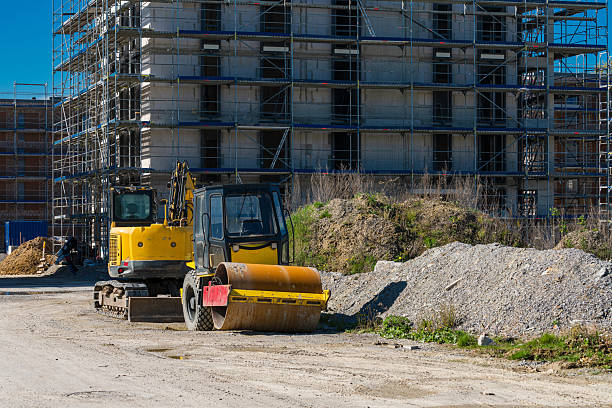 Construction activity is underway at a site featuring scaffolding and multiple heavy machines. A yellow roller and an excavator are parked near piles of gravel and soil under a clear blue sky.